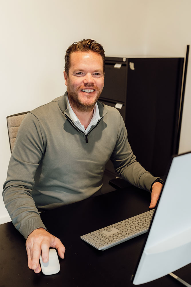 Simon Hulsman, financieel adviseur bij Meierijstad Hypotheken, glimlacht achter zijn bureau terwijl hij werkt op een computer in een moderne kantooromgeving. Foto gebruikt op de pagina over zijn hypotheekadviesdiensten binnen het makelaarskantoor.